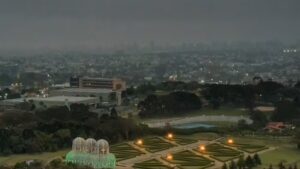 Cidade sob céu nublado com iluminação de ruas e jardim geométrico em primeiro plano