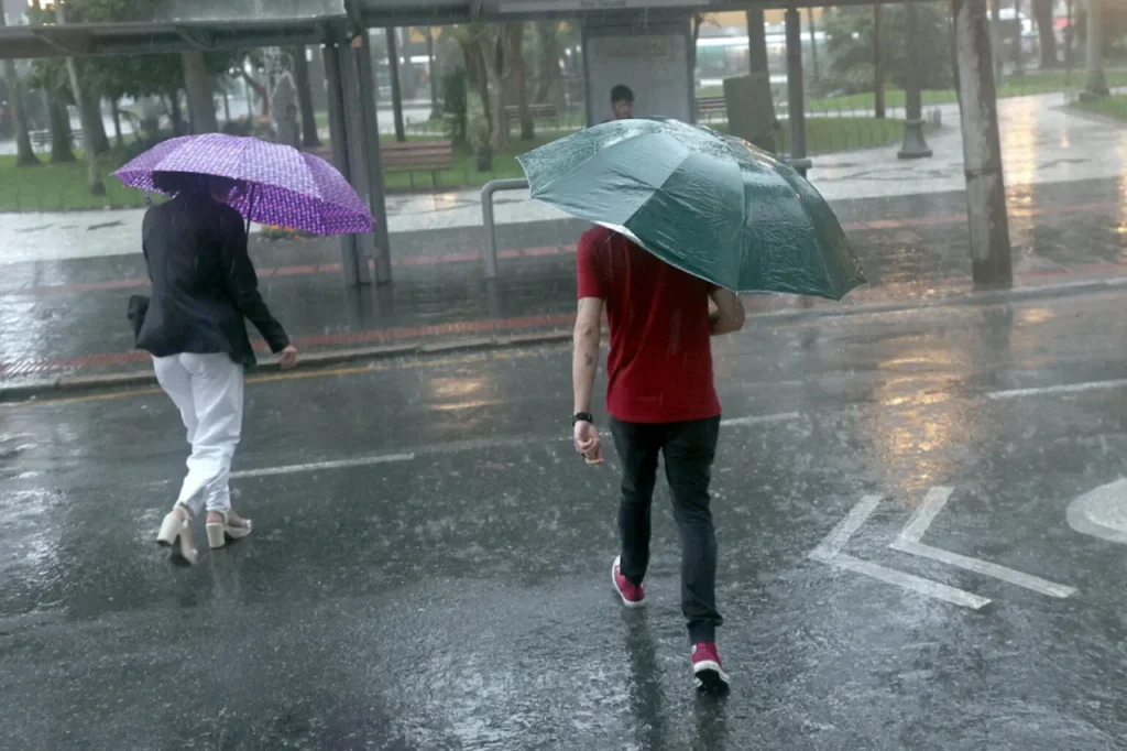 Duas pessoas com guarda-chuvas caminham em rua alagada durante forte chuva em área urbana