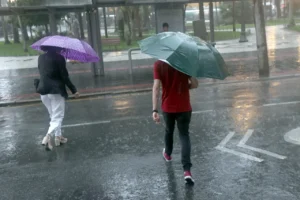 Duas pessoas com guarda-chuvas caminham em rua alagada durante forte chuva em área urbana