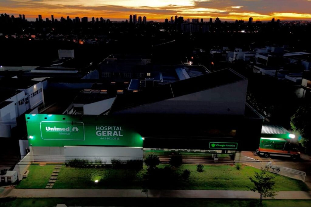 Vista aérea noturna de um hospital com letreiro iluminado e edifícios ao fundo sob céu colorido