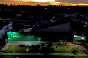 Vista aérea noturna de um hospital com letreiro iluminado e edifícios ao fundo sob céu colorido
