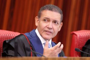 Um homem de toga e terno sentado em cadeira vermelha, falando com gesto de mão em ambiente formal