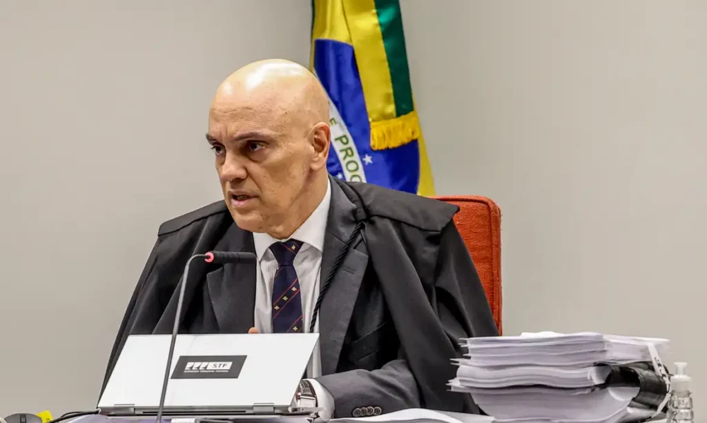 Um homem de toga sentado em uma mesa com microfone e documentos, diante de uma bandeira