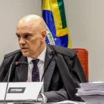 Um homem de toga sentado em uma mesa com microfone e documentos, diante de uma bandeira
