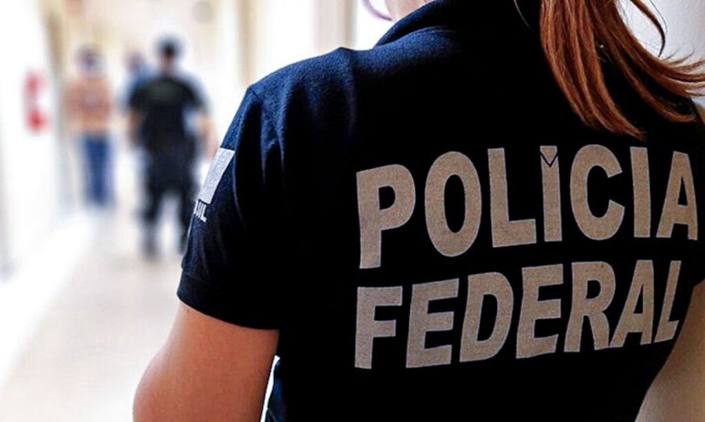 Pessoa com camisa que diz "POLÍCIA FEDERAL" em corredor com outras pessoas ao fundo