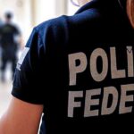 Pessoa com camisa que diz "POLÍCIA FEDERAL" em corredor com outras pessoas ao fundo