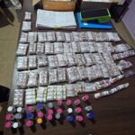 Mesa com pacotes embalados, frascos coloridos, documentos e equipamentos, ao lado de distintivo da Polícia Civil