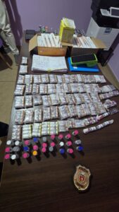 Mesa com pacotes embalados, frascos coloridos, documentos e equipamentos, ao lado de distintivo da Polícia Civil