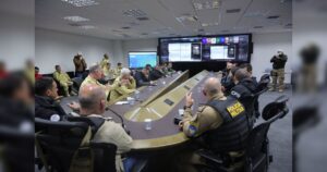 Um grupo de pessoas em uniformes militares e civis reúne-se em sala com painéis eletrônicos para discussão estratégica