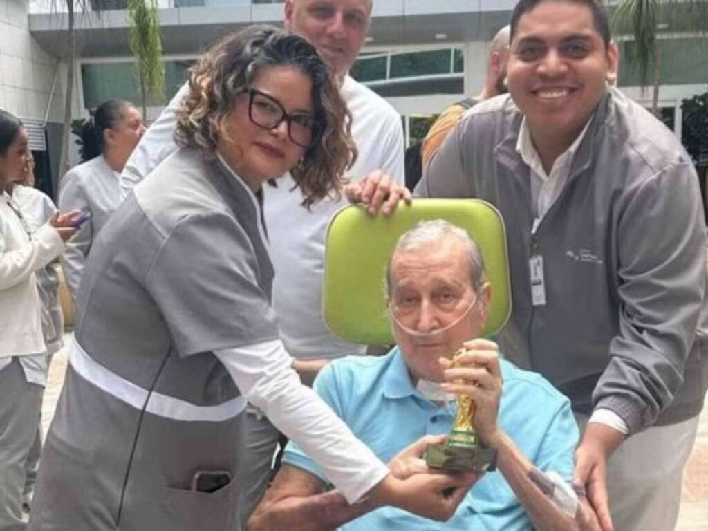 Um homem idoso com tubo de oxigênio sentado em cadeira é ajudado por profissionais e segura uma pequena estátua dourada ao sair de um prédio