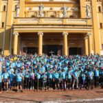 Um grupo de pessoas em camisetas azuis posa em frente a uma igreja amarela com colunas e estátuas