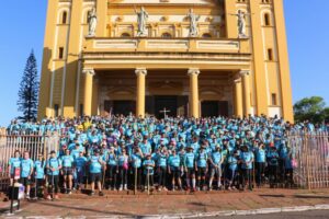 Um grupo de pessoas em camisetas azuis posa em frente a uma igreja amarela com colunas e estátuas