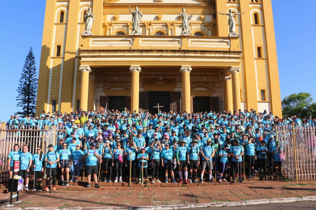Um grupo de pessoas em camisetas azuis posa em frente a uma igreja amarela com colunas e estátuas