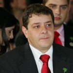 Um homem de terno e gravata vermelha em evento com outras pessoas ao fundo
