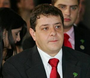 Um homem de terno e gravata vermelha em evento com outras pessoas ao fundo