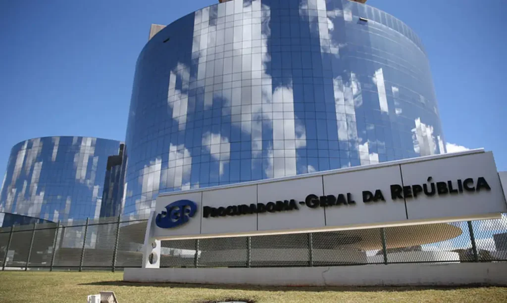 Edifício com fachada de vidro refletindo o céu e nuvens, com placa da Procuradoria-Geral da República à frente