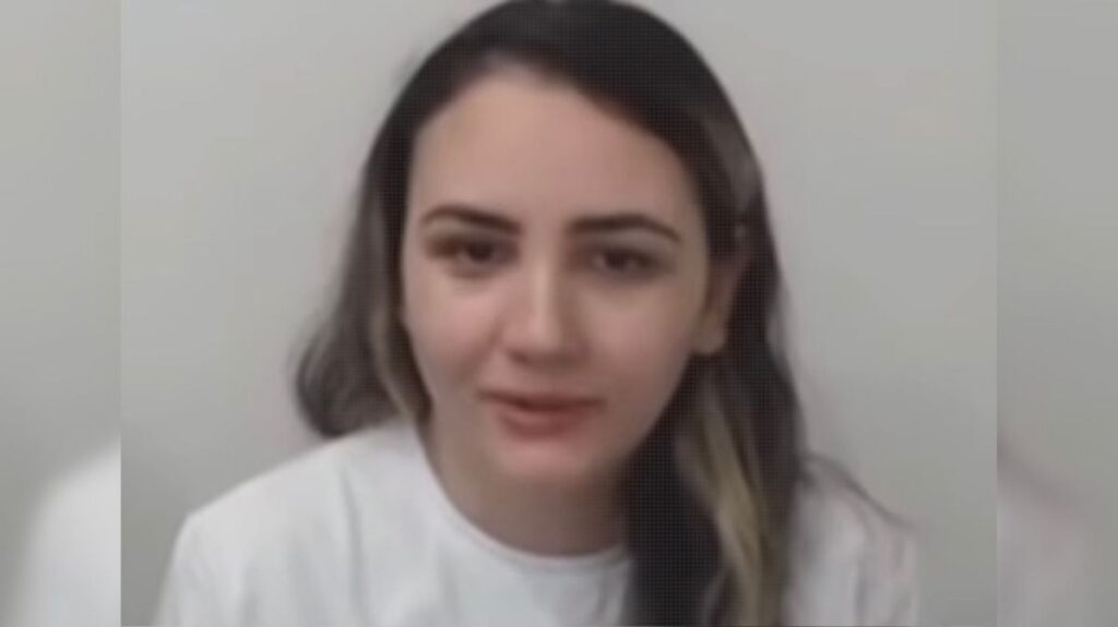 Uma mulher com cabelo escuro e mechas claras veste camiseta branca e fala em frente a fundo claro