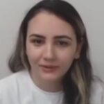 Uma mulher com cabelo escuro e mechas claras veste camiseta branca e fala em frente a fundo claro