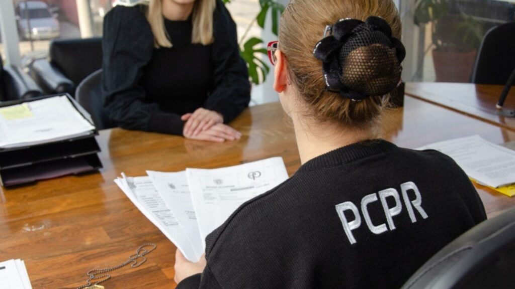 Uma pessoa com camiseta que exibe as letras PCPR sentada à mesa com documentos em frente a outra pessoa