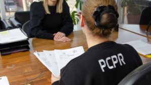Uma pessoa com camiseta que exibe as letras PCPR sentada à mesa com documentos em frente a outra pessoa
