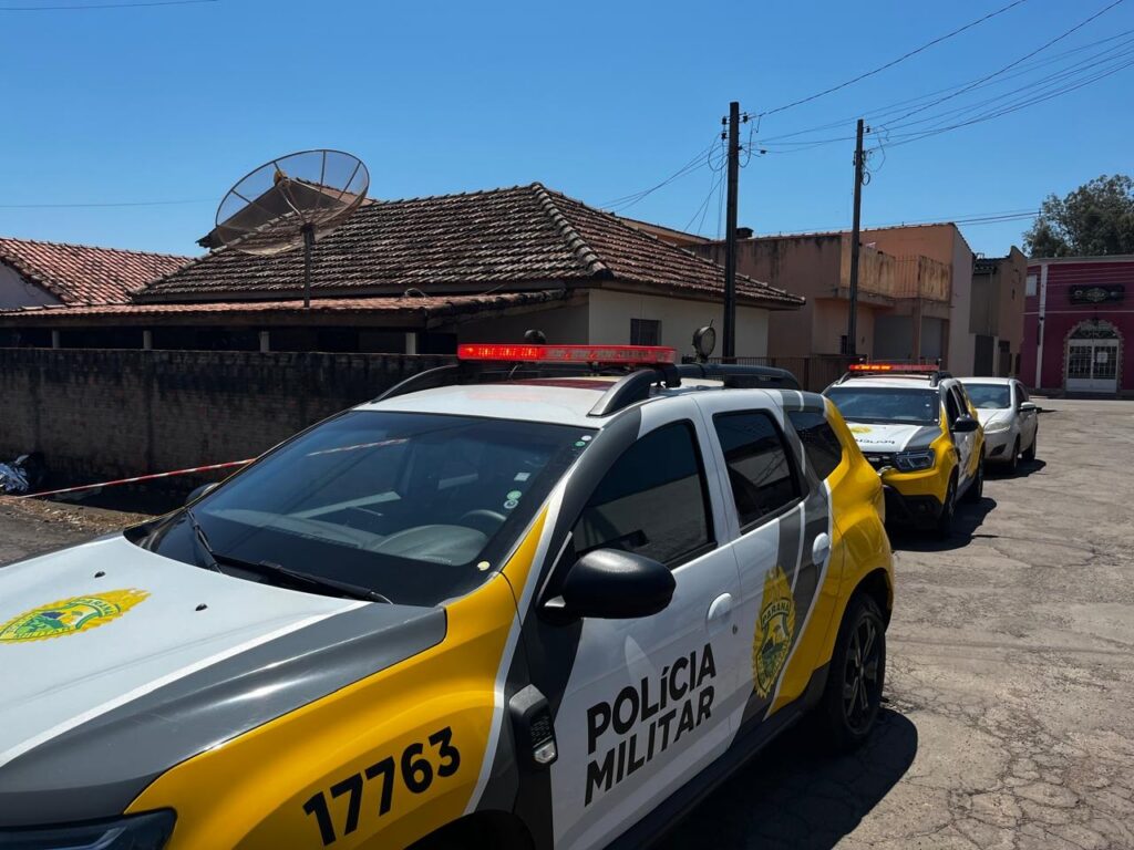 Veículos da Polícia Militar estacionados em rua com faixa de isolamento e casas ao fundo