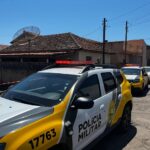 Veículos da Polícia Militar estacionados em rua com faixa de isolamento e casas ao fundo
