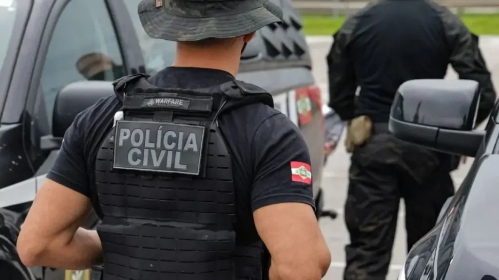 Um homem com colete da Polícia Civil em pé ao lado de veículos em uma via movimentada