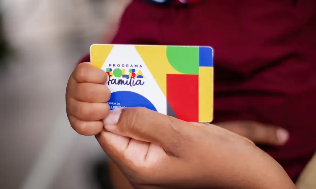 Mãos seguram cartão do Programa Bolsa Família com design colorido e logotipo visível