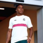 Um jovem com camisa branca do Real Madrid e detalhe rosa posa em ambiente interno