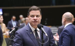 Um homem de terno fala em microfone em ambiente com outras pessoas ao fundo