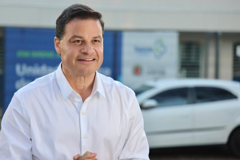Um homem de camisa branca posa em frente a um prédio com veículos ao fundo