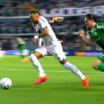 Um jogador de branco com a bola é pressionado por dois adversários em campo de futebol