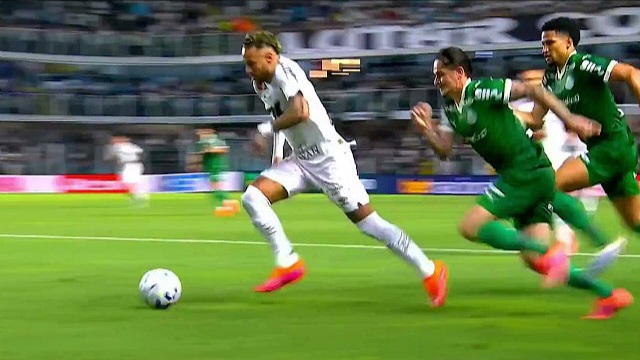 Um jogador de branco com a bola é pressionado por dois adversários em campo de futebol
