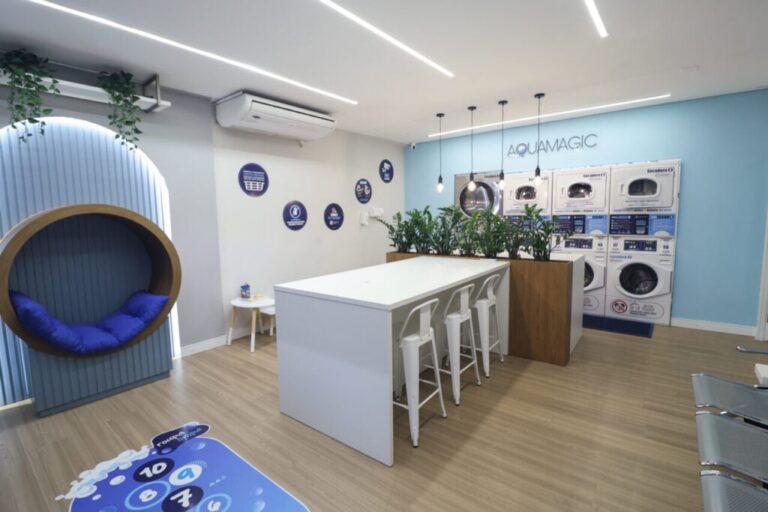 Espaço interno com máquinas de lavar, mesa com banquetas e decoração moderna em tons de azul e branco