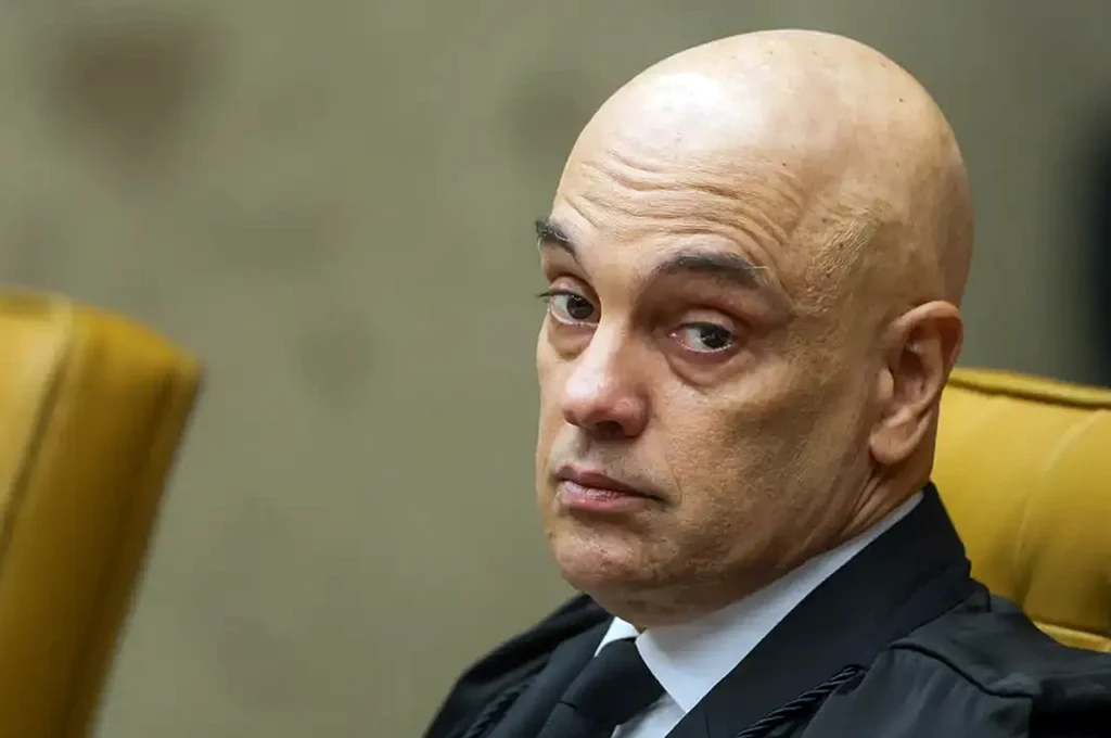 Um homem de terno e capa preta sentado em cadeira amarela em ambiente formal