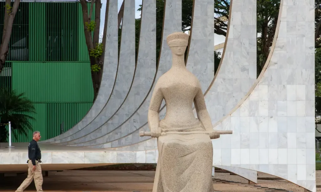 Estátua de mulher com cinto e olhos vendados em frente a estrutura de mármore com arcos curvos, ao fundo um homem caminha perto de parede verde