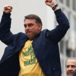 Um homem com camiseta amarela e jaqueta escura levanta os punhos em frente a um prédio