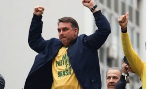 Um homem com camiseta amarela e jaqueta escura levanta os punhos em frente a um prédio