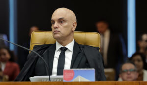 Um homem de toga sentado em cadeira de couro em plenário com microfone e documentos à frente