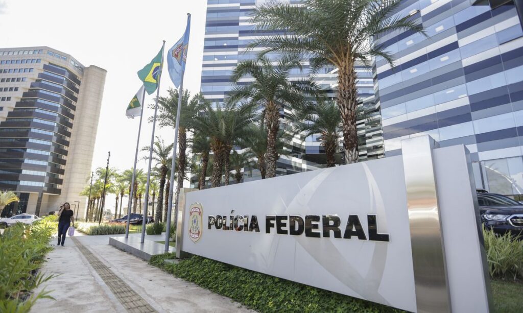 Fachada da Polícia Federal com prédio moderno, palmeiras e bandeiras ao fundo