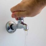Uma mão abre uma torneira com pouca água saindo em um ambiente interno
