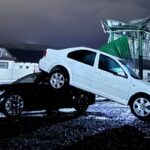 Carro branco sobre carro preto em área alagada com estrutura danificada ao fundo