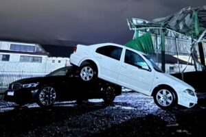 Carro branco sobre carro preto em área alagada com estrutura danificada ao fundo