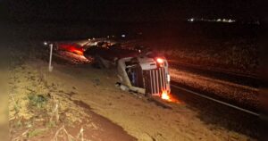 Bitrem tombado à noite em acostamento de rodovia, com luzes acesas e carga espalhada ao redor