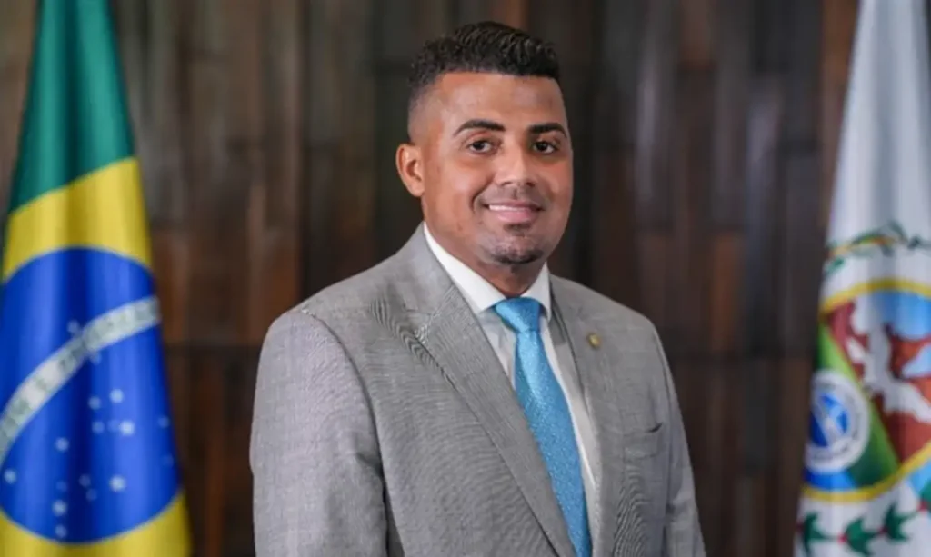 Um homem de terno cinza e gravata azul posa entre duas bandeiras em ambiente interno com painel de madeira