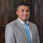 Um homem de terno cinza e gravata azul posa entre duas bandeiras em ambiente interno com painel de madeira