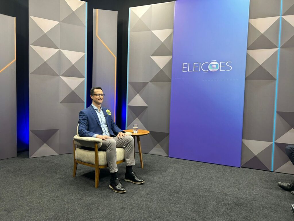 Um homem sentado em cadeira durante evento com painel que diz "ELEIÇÕES" ao fundo