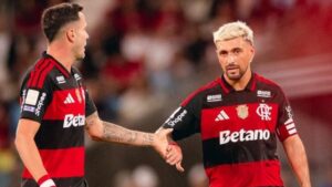Dois jogadores de camisa preta e vermelha se cumprimentam com aperto de mão em campo de futebol