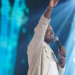 Um homem canta no palco com microfone na mão e braço erguido sob luzes de show