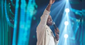 Um homem canta no palco com microfone na mão e braço erguido sob luzes de show